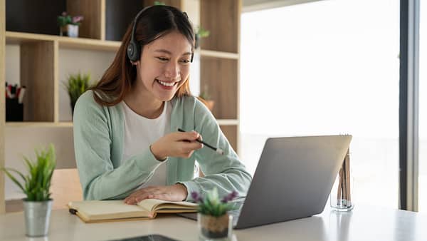 Woman sitting in an office and wearing headphones smiles while pointing a pencil at her laptop